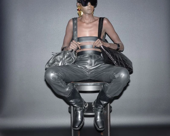 Model seated on a chair wearing dark leather trousers and boots while holding two handbags against a grey gradient background