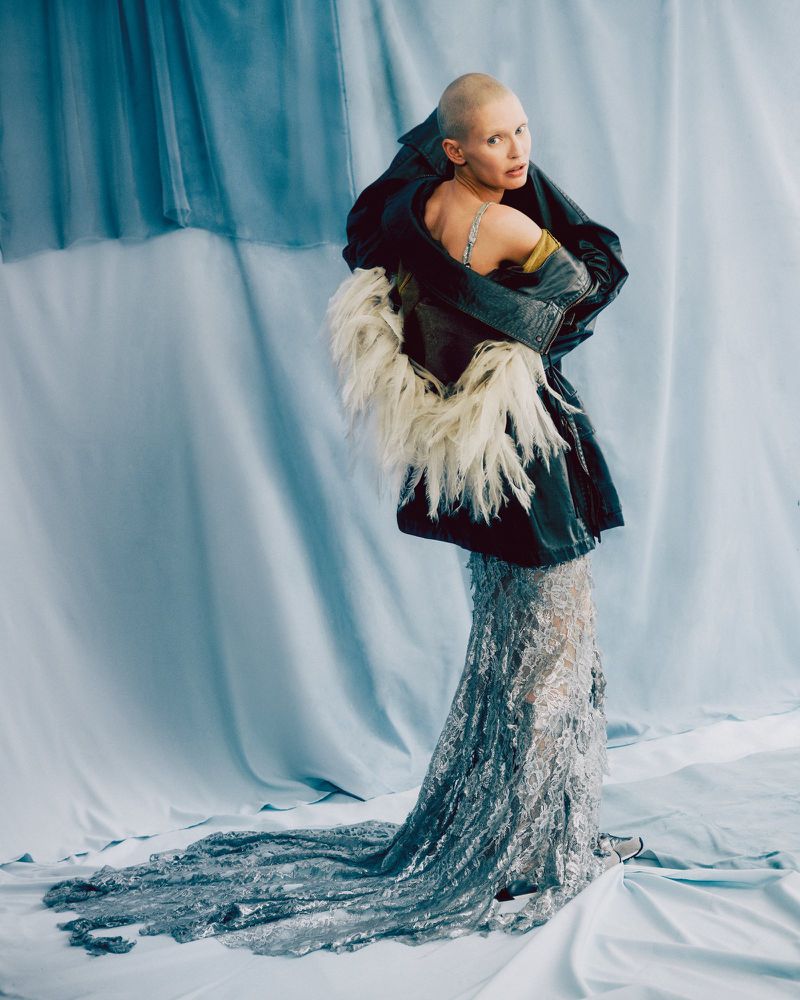 A woman with a shaved head in a dark jacket with a feather collar poses in a silver glittery gown against a light blue draped background.
