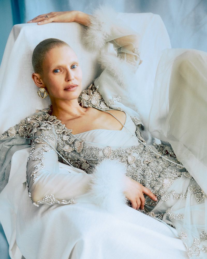Elegant woman in ornate dress reclines against soft fabric, showcasing detailed embellishments and sophisticated fashion in a soft lighting.
