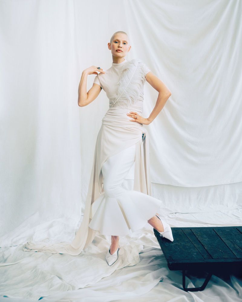 A person with a shaved head poses in a draped white dress, featuring white shoes and a dark table, set against a white fabric background.
