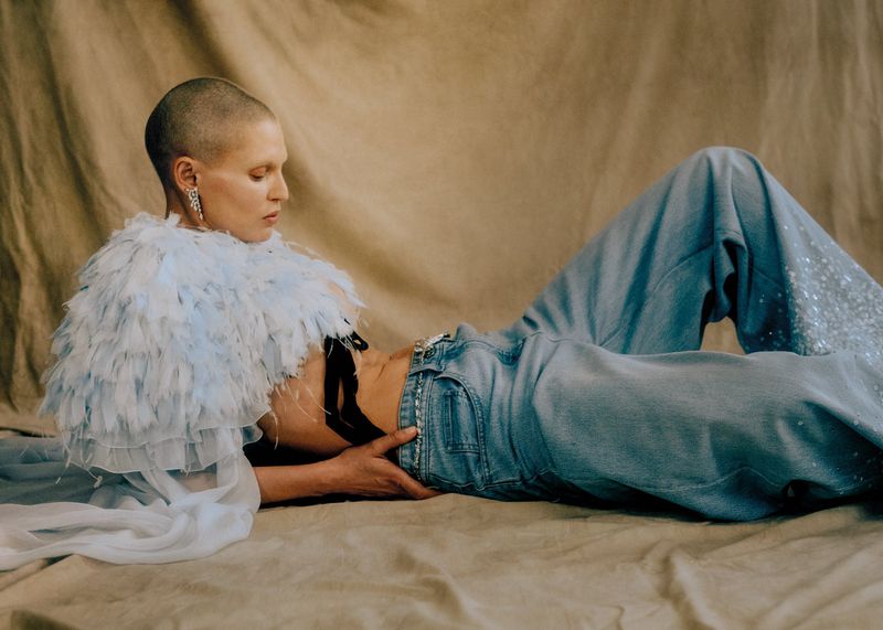 A woman with a shaved head and light jeans lies on a tan backdrop, wearing a feathery light blue top, captured in a studio portrait.