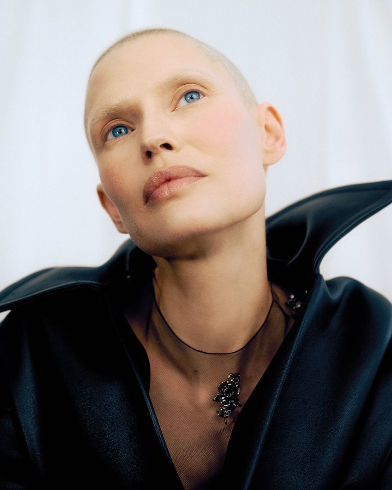 Woman with shaved head wearing a black top and statement necklace looking up against a white background in a studio portrait.