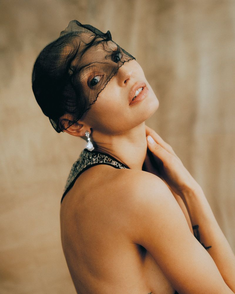 Portrait of woman with sheer black face covering, elegant silver earrings, and hand to her neck, against a tan background.