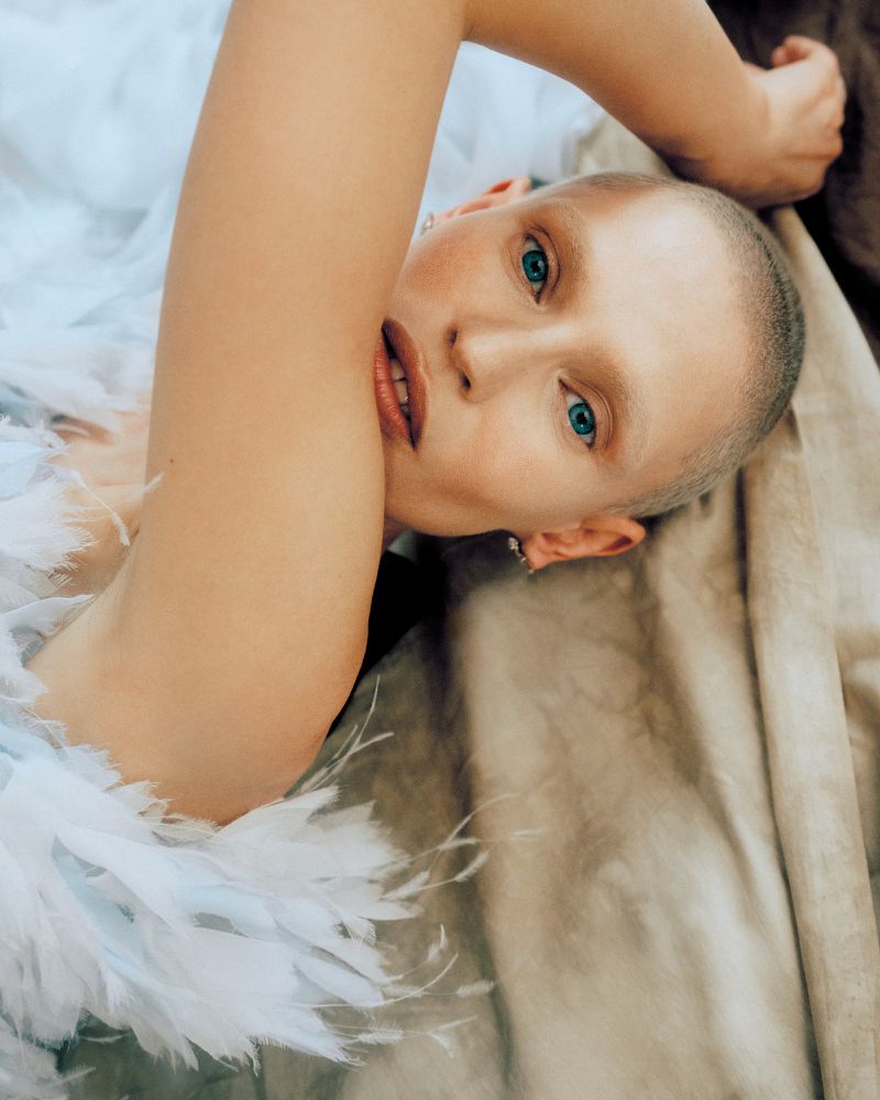 Adult woman with short hair lies down in feathered white gown. She has fair skin, blue eyes, and her head is tilted.