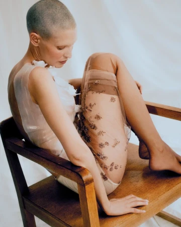 Studio portrait of a bald, fair-skinned woman in a sheer dress sitting on a wooden chair against a clean, white background.