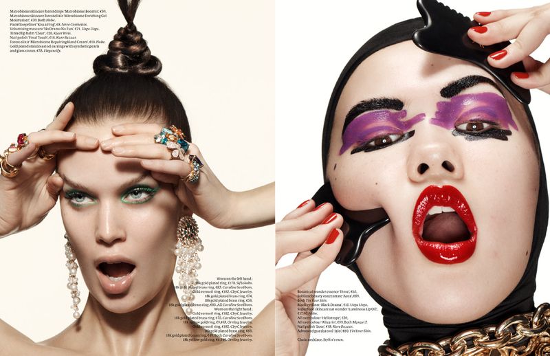 Close-up of two women with artistic makeup, red lipstick, multiple rings, a headscarf, and statement earrings in a studio.