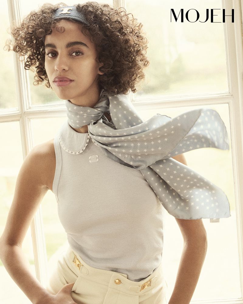 Fashion portrait of an adult woman with curly hair wearing a sleeveless top and a patterned scarf near a window with a garden view.