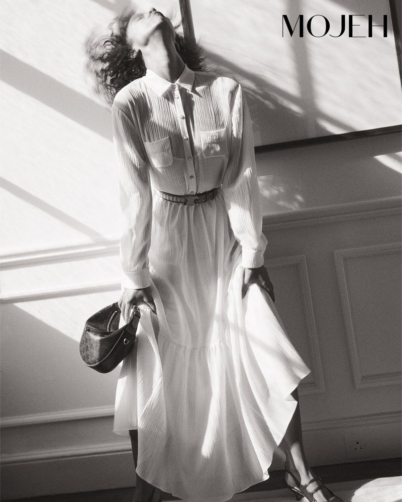 Monochrome shot of a stylish adult woman in a white dress, holding a purse, with light casting shadows on the wall behind her.