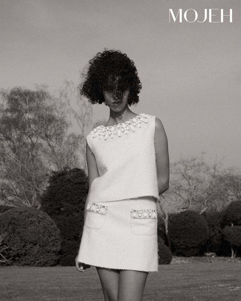 Monochrome fashion shot: woman in a sleeveless embellished top and mini skirt stands in front of trees and a bright sky.