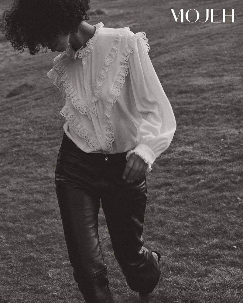Monochrome shot of a woman with curly hair wearing a ruffled white blouse and leather pants, posing outdoors in a grassy field.