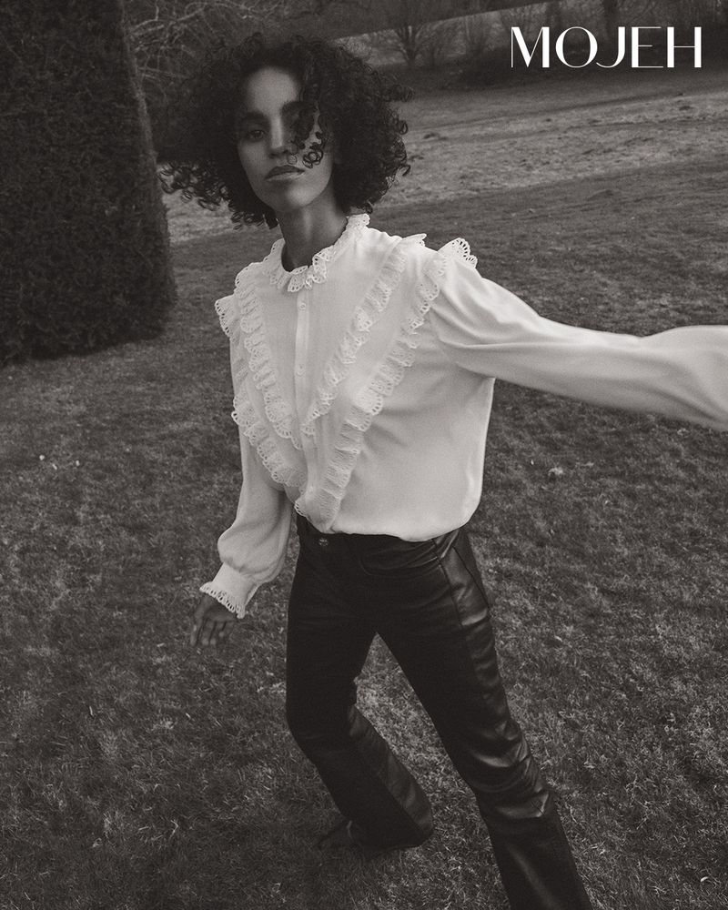 Monochrome fashion shot: a slender model with curly hair wears a frilly white blouse and black leather pants outdoors in a grassy field.