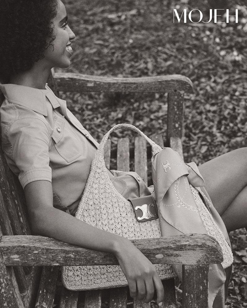 A woman sits on a wooden bench with a woven handbag in a black and white photograph. She is smiling with trees in background.