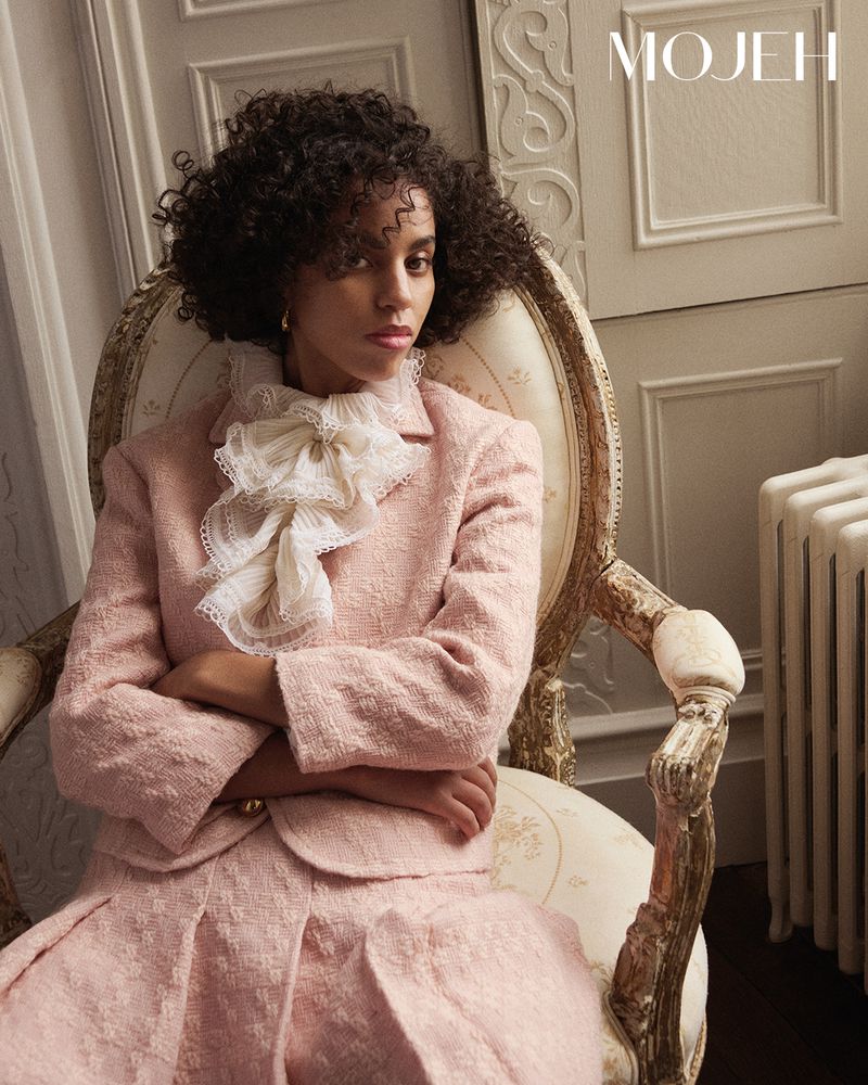 A woman with curly hair sits in an ornate chair, wearing a light pink tweed suit with a large lace collar and the MOJEH magazine logo.