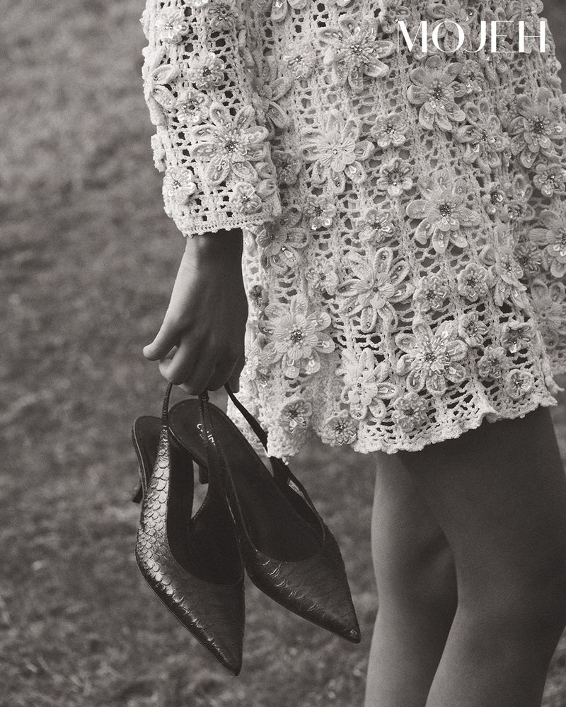 Cropped view of a woman wearing a flower-embellished dress, holding a pair of slingback pumps in a black and white outdoor shot.