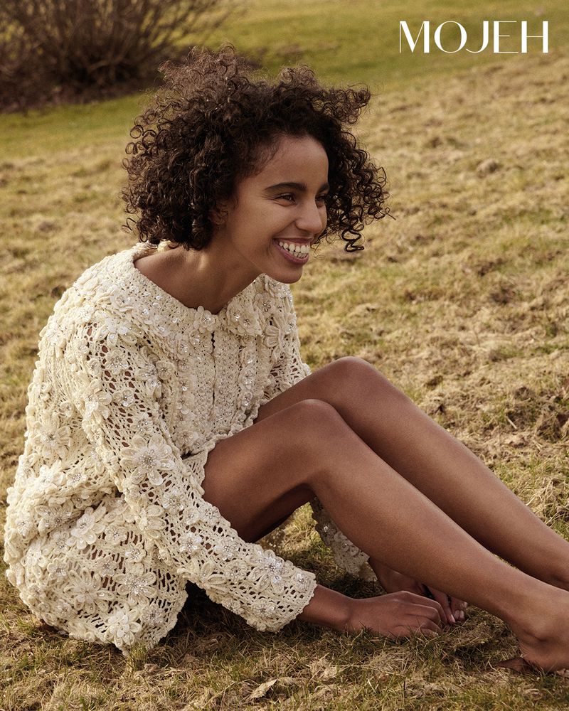 Smiling model with curly hair wearing a cream lace dress sitting outdoors on grassy terrain.
