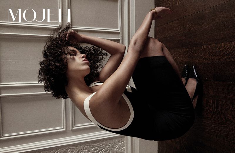 Woman in black dress and high heels poses against a paneled wall and wood floor for MOJEH magazine fashion editorial.