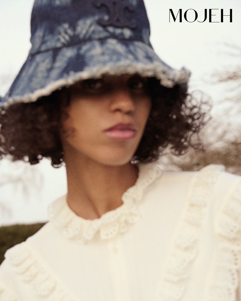 Close-up portrait of a woman with curly hair wearing a tie-dye bucket hat and white frilly top, set against a blurred outdoor background.