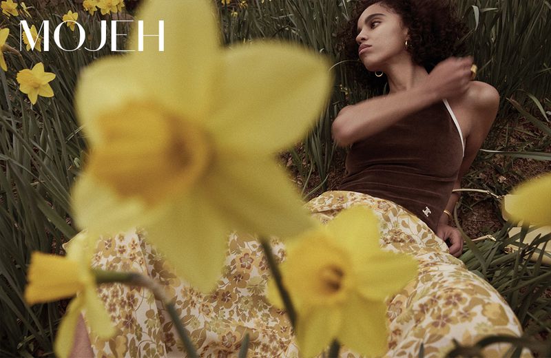 Fashion editorial image of a woman with curly hair in a brown top and patterned pants lying amongst yellow daffodils and green foliage.