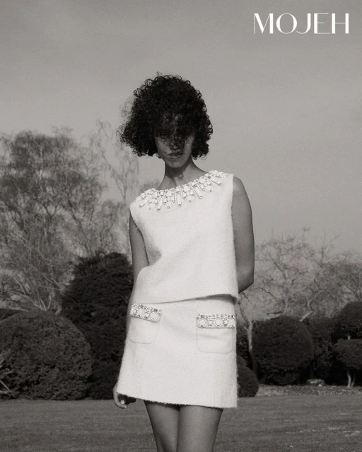 Model with dark curly hair wearing an embellished white tweed skirt suit outdoors in a garden for Mojeh Magazine