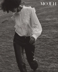 Model with dark curly hair wearing a white ruffled blouse and dark trousers walking outdoors for Mojeh Magazine