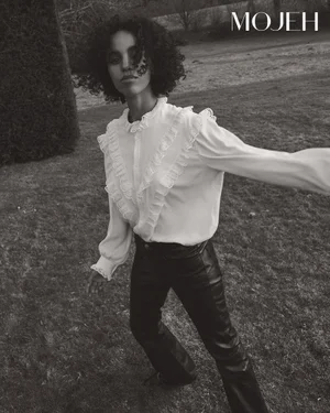 Model with curly hair in a white ruffled blouse and dark pants outdoors on a grassy lawn for Mojeh Magazine editorial.