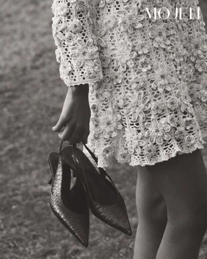 Model wearing a white crochet dress holding dark slingback heels in a black and white fashion editorial for Mojeh Magazine