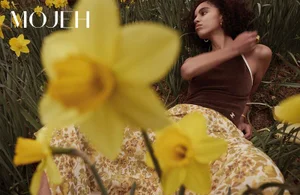 Model with dark curly hair reclining among yellow daffodils wearing a brown top and patterned skirt for Mojeh Magazine