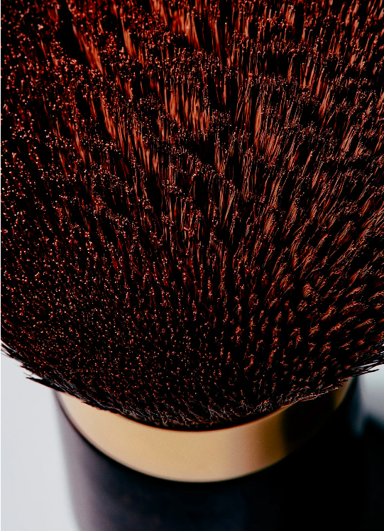 Closeup of a makeup brush with dark brown bristles, gold band, and dark handle, showcasing texture, detail, and color in a beauty context.