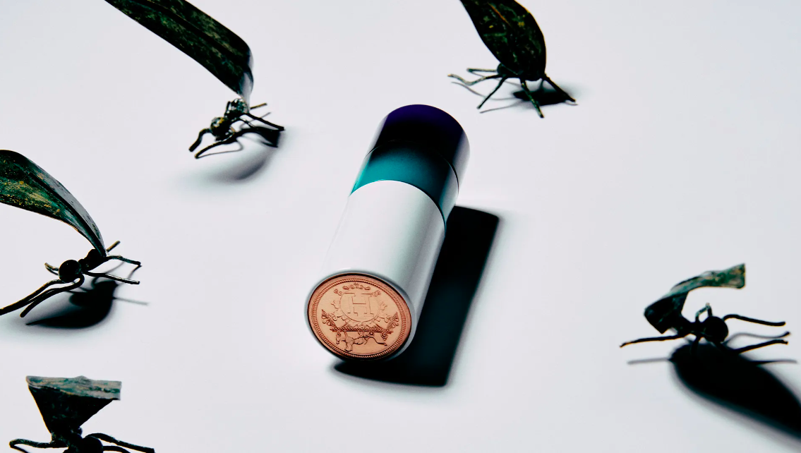 Cylindrical container with a round emblem surrounded by dark metal insect sculptures on a bright white surface.