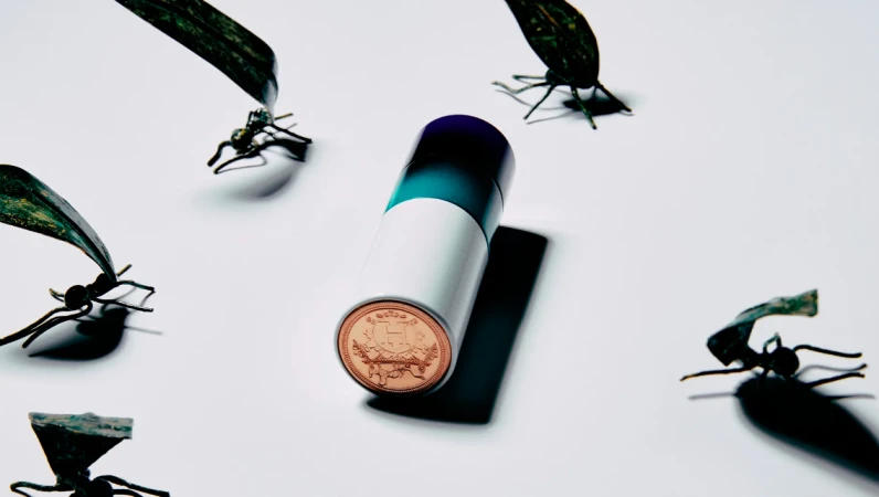 Cylindrical container with a round emblem surrounded by dark metal insect sculptures on a bright white surface.