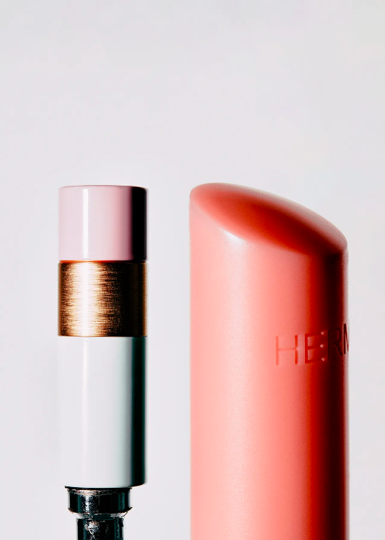 Close-up studio shot of two lipstick tubes, one with a pink cap, gold band, and silver base, the other peach, against a white backdrop.