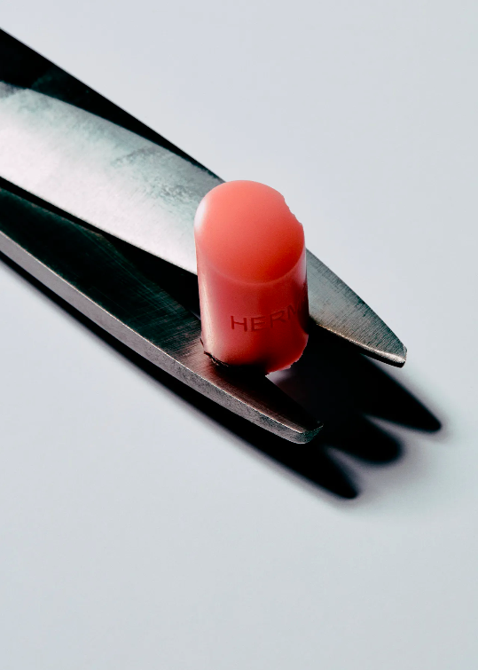 Close-up studio shot of peach-colored lipstick bullet held by the blades of a pair of stainless steel scissors on a white surface.