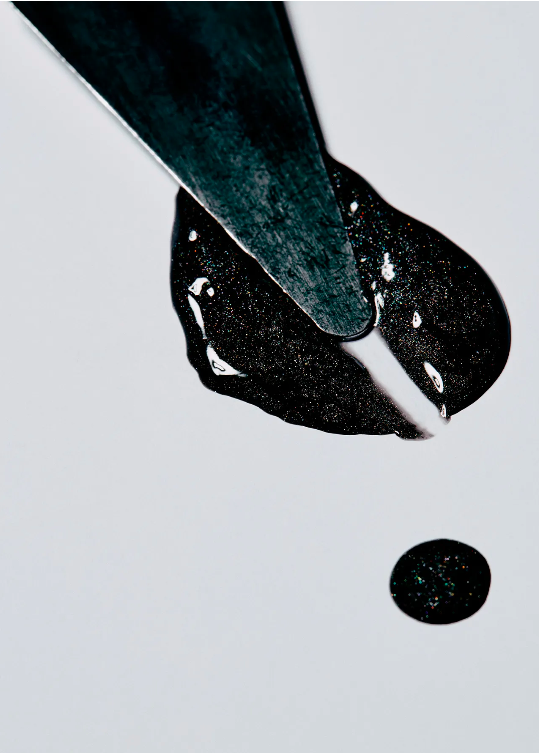 Close-up of a black viscous liquid being spread by a silver metal spatula on a white surface; studio shot with minimal light.