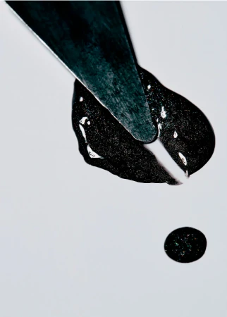 Close-up of a black viscous liquid being spread by a silver metal spatula on a white surface; studio shot with minimal light.
