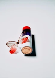Lipstick tube standing upright next to translucent reddish insect wings on a white seamless background
