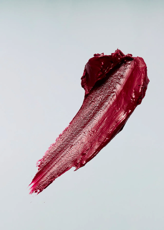 Close-up of a smear of dark red lipstick against a light gray background, highlighting texture and color for beauty product promotion.