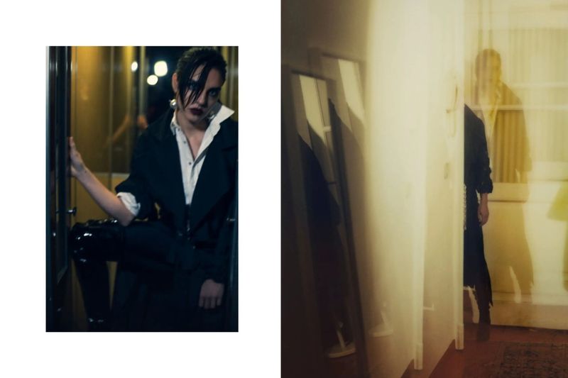Adult posing indoors in a black coat and white shirt leaning in a doorway with a white wall and shadowy figure in the background.