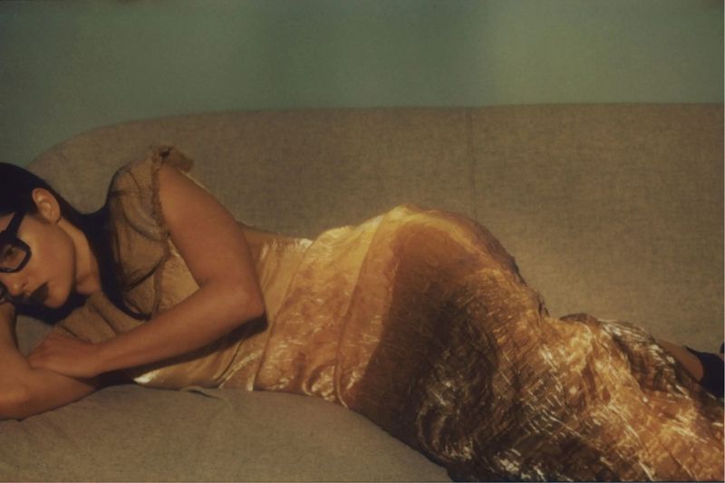 Adult woman in gold dress lying on a light-colored couch, wearing eyeglasses, indoors with soft, diffused lighting, and a minimal background.
