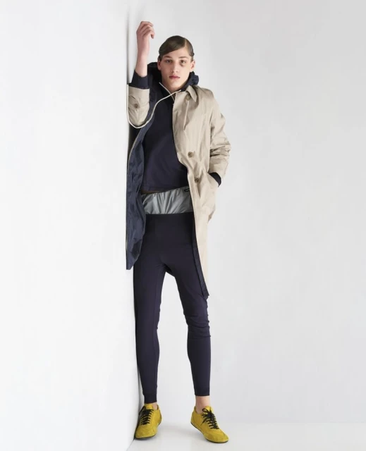Full shot of a person leaning against a white wall. They wear a tan trench coat over a navy blue hoodie with pants and yellow sneakers.