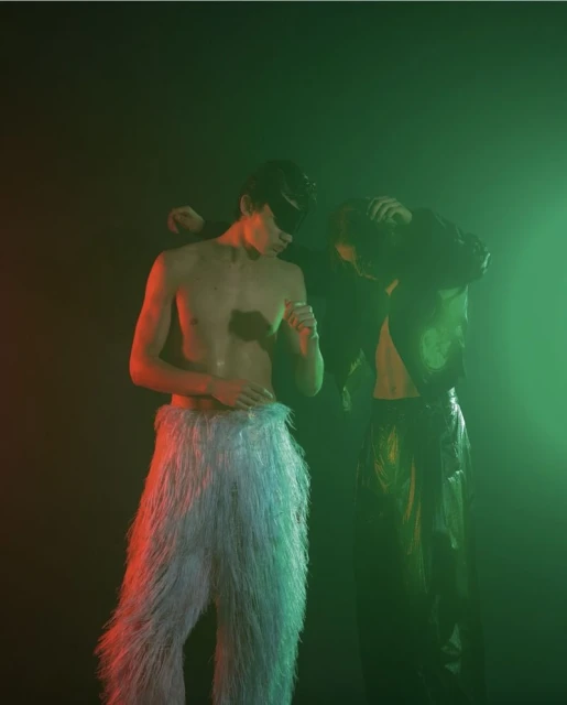 Two young adults in a dark, hazy setting. One wears furry pants and a cap, while the other is in a shiny outfit.