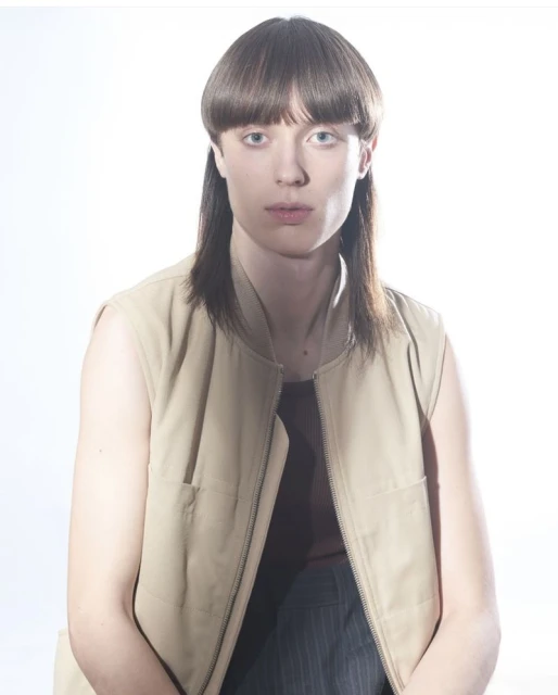 Close-up studio portrait of a person with bowl cut hairstyle and vest. The person has fair skin and blue eyes against a white backdrop.