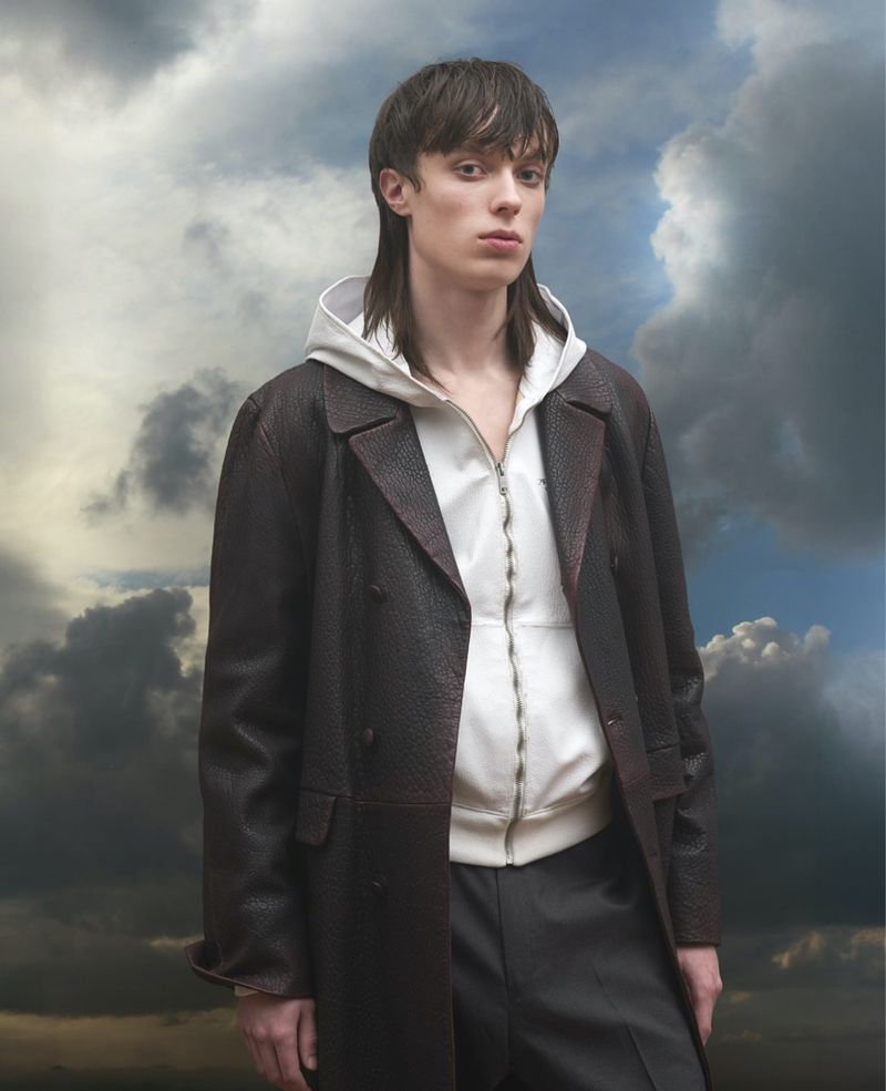 Portrait of a young adult man in a brown coat and white hoodie against a cloudy sky, striking a confident pose with a serious facial expression.