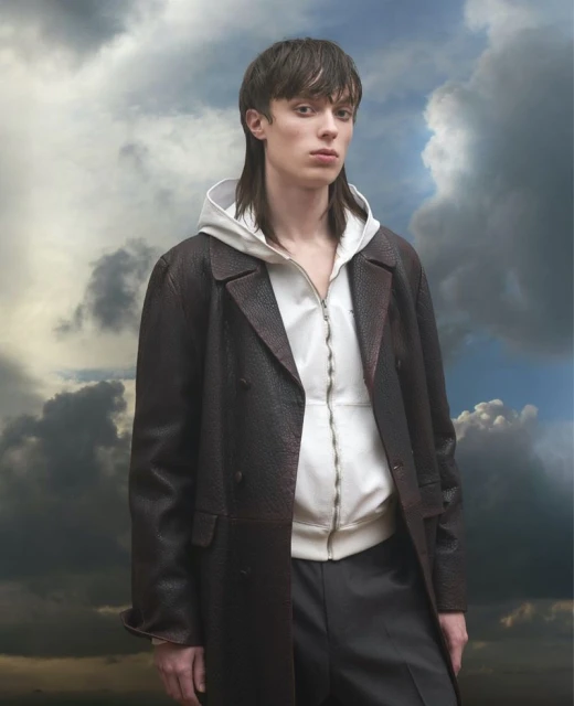 Portrait of a young adult man in a brown coat and white hoodie against a cloudy sky, striking a confident pose with a serious facial expression.