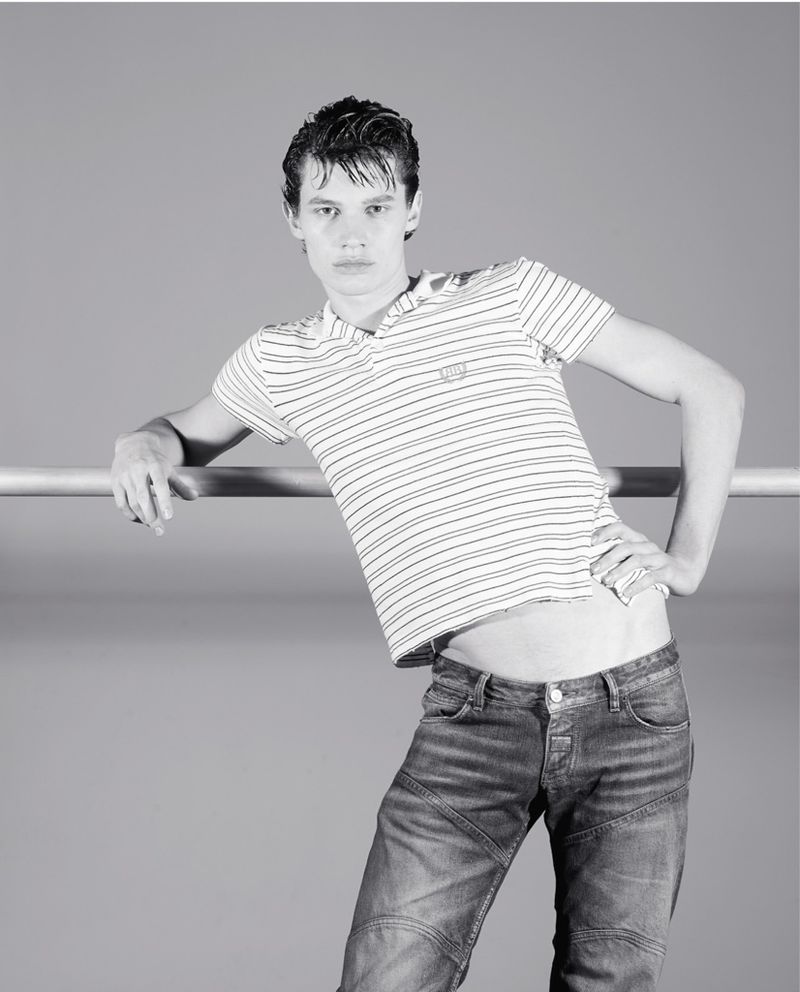 A male fashion model in a striped shirt and blue jeans leans against a metal bar in a black and white studio portrait.