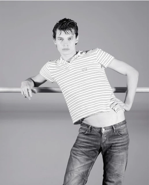 A male fashion model in a striped shirt and blue jeans leans against a metal bar in a black and white studio portrait.