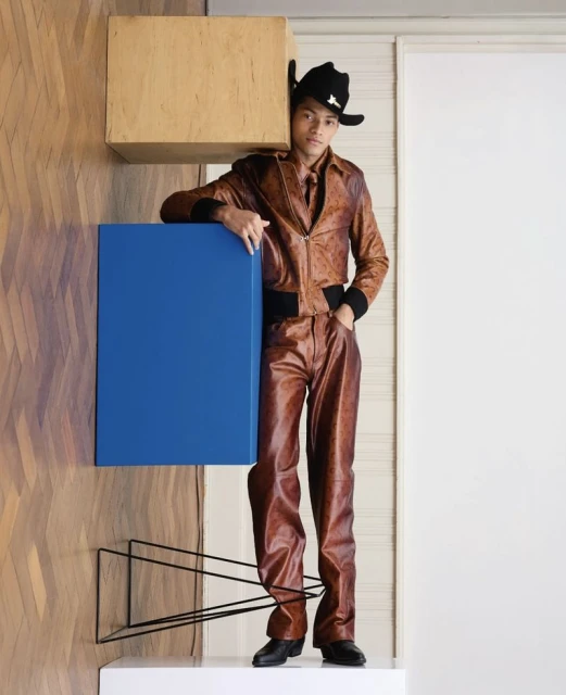 Man in brown leather jacket and pants with black cowboy hat, posing between wood and blue cubes in a minimalist indoor setting.