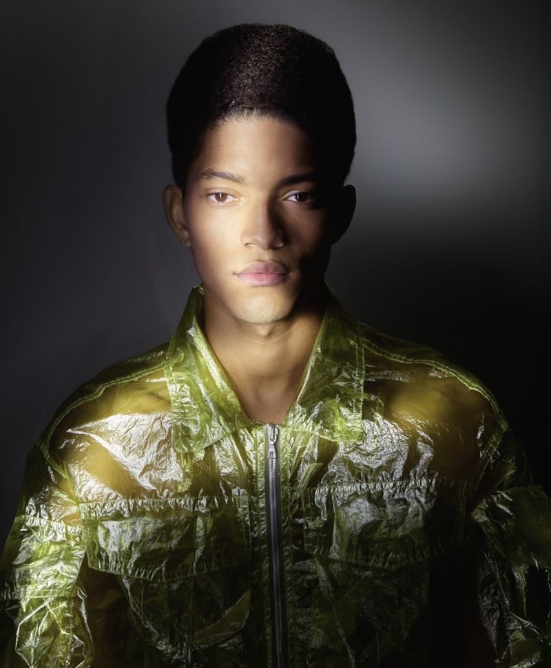 Portrait of a dark-skinned young adult male in a translucent yellow jacket against a dark gray background with dramatic lighting.