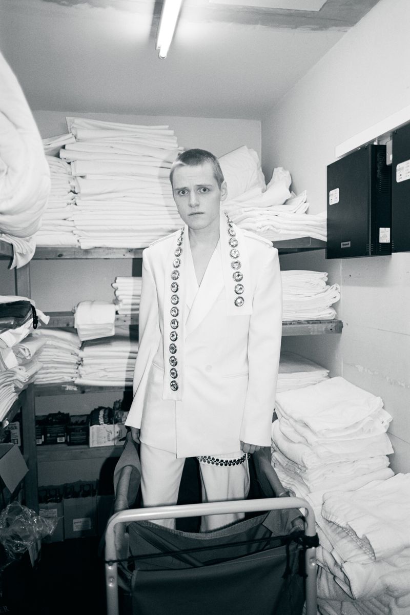 Monochrome shot of a person in a white suit standing in a metal cart surrounded by stacks of folded white linens in a storage room.
