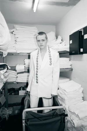 Monochrome shot of a person in a white suit standing in a metal cart surrounded by stacks of folded white linens in a storage room.