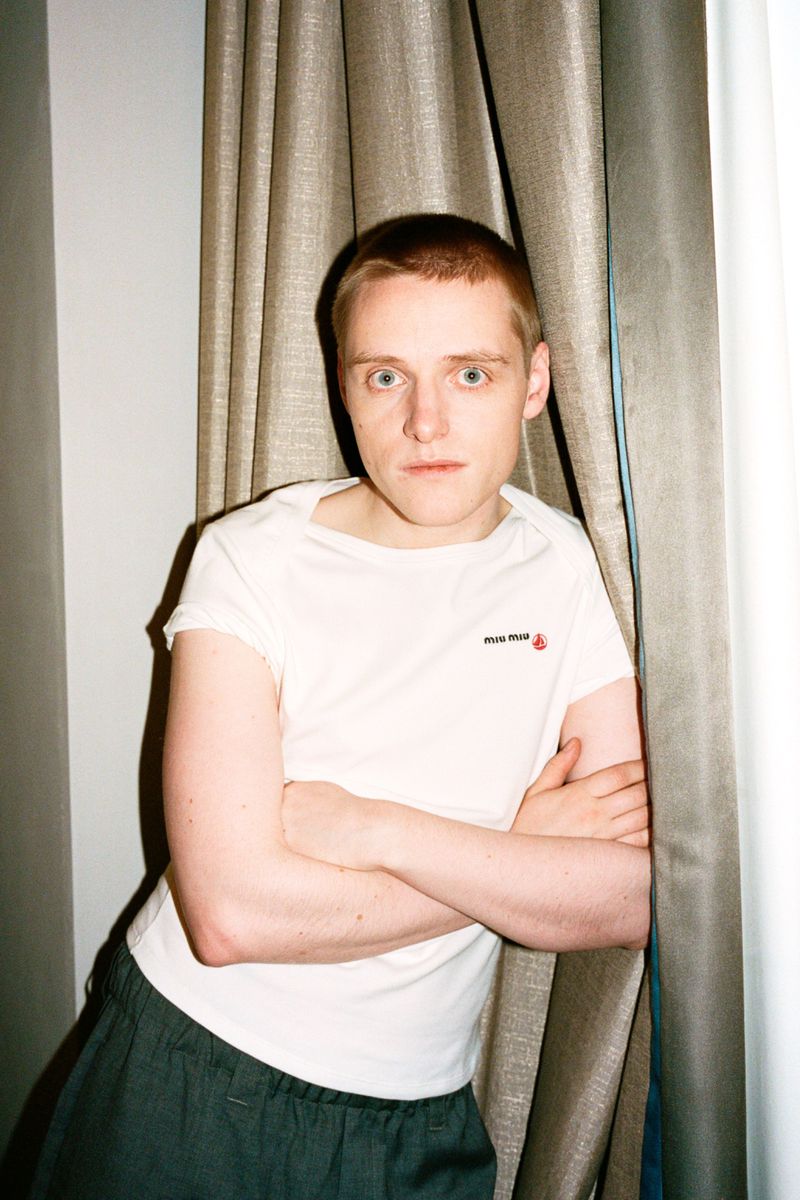 Young fair-skinned man in white tee with crossed arms stands next to a draped curtain indoors, portrait orientation.